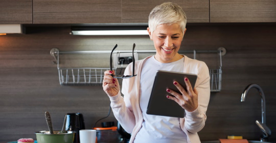 Middle-aged woman removing her glasses and smiling at her tablet device in a kitchen
